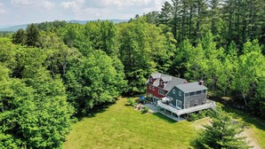 A two-story house with a large deck and a covered patio, surrounded by dense forest. - SPLISH SPLASH! Explore Vermont! (Londonderry)