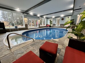 Une piscine intérieure dotée d’un sol en pierre, de sièges rembourrés rouges et de grandes fenêtres offrant une vue sur les arbres.
