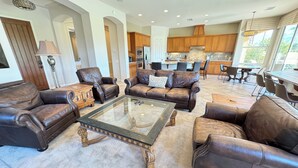 A spacious living room with leather sofas, a glass coffee table, and a kitchen in the background.