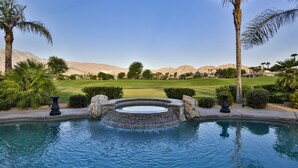 A swimming pool with a waterfall feature, surrounded by palm trees and a view of a golf course and mountains.