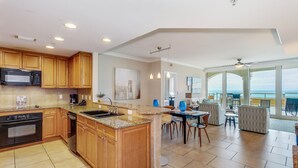 A spacious kitchen with wooden cabinets, a granite countertop, and a large island. There is a dining area with a table and chairs, and a view of the ocean through large windows. - P5-0503 Stunning Gulf Front Terrace Sunset Views! (Pensacola Beach)