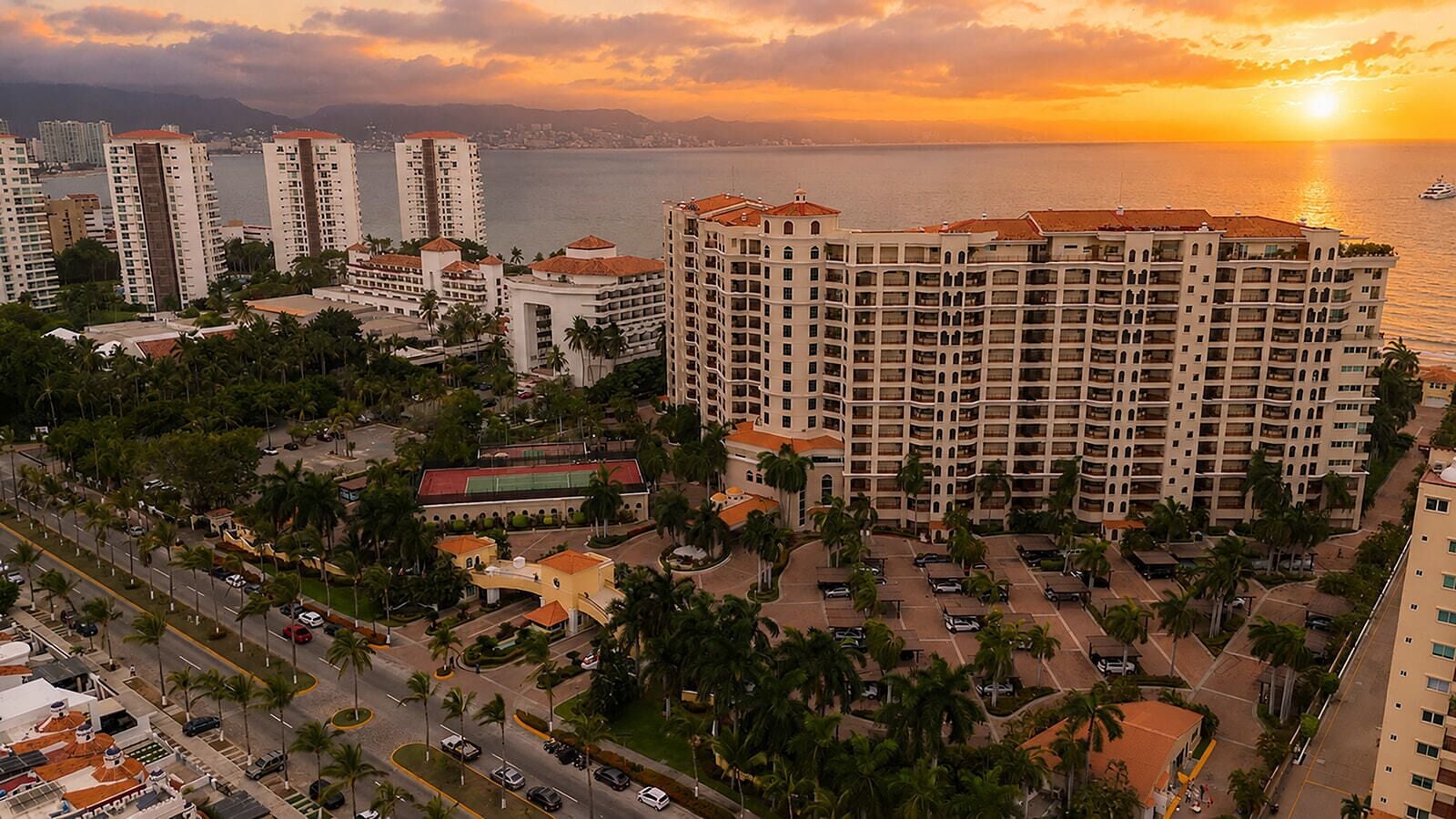 A high-rise hotel with multiple floors, a parking area, and a view of the sea at sunset.