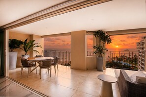 Un balcon offrant une vue sur le coucher de soleil sur l’océan, un coin repas avec une table et des chaises, et des plantes en pot.