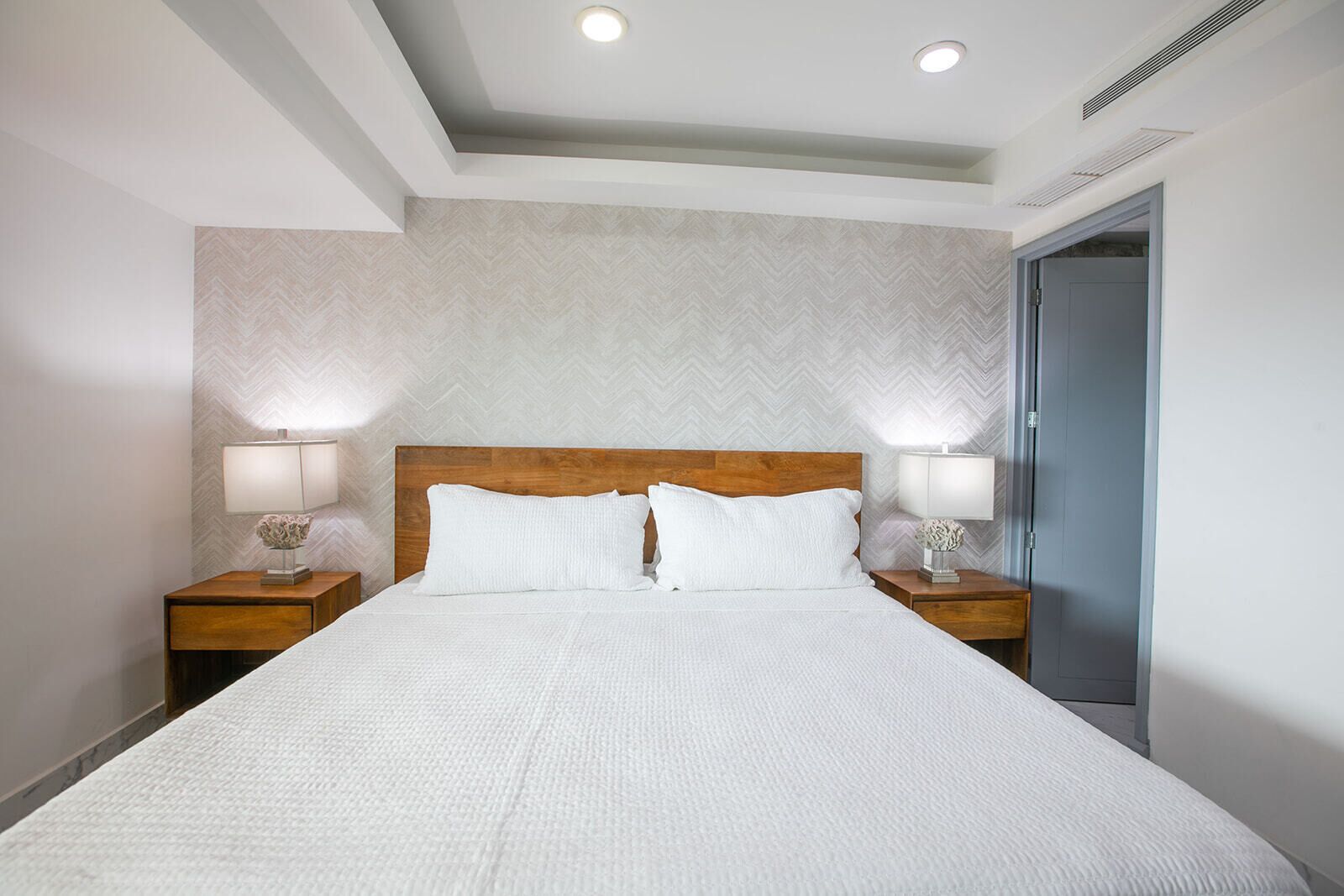 Habitación de hotel con una cama grande, dos mesitas de noche con lámparas, un cabecero de madera y un papel tapiz estampado.