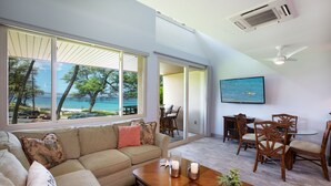 Een woonkamer met een groot raam dat uitkijkt op een strand, een houten eettafel en een plafondventilator.