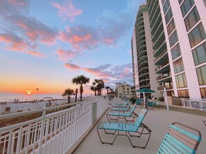 A beachfront hotel with a balcony overlooking the ocean, featuring rows of lounge chairs and a white railing.