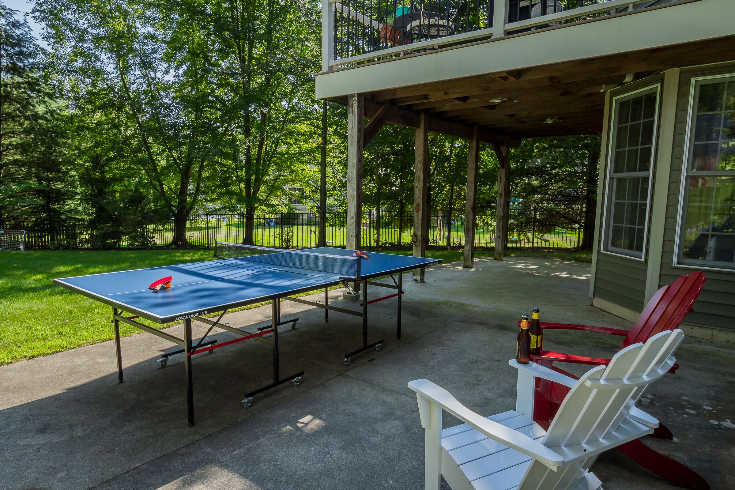 Une table de ping-pong installée sur une terrasse en béton, avec une chaise Adirondack rouge et blanche et deux bouteilles de bière.