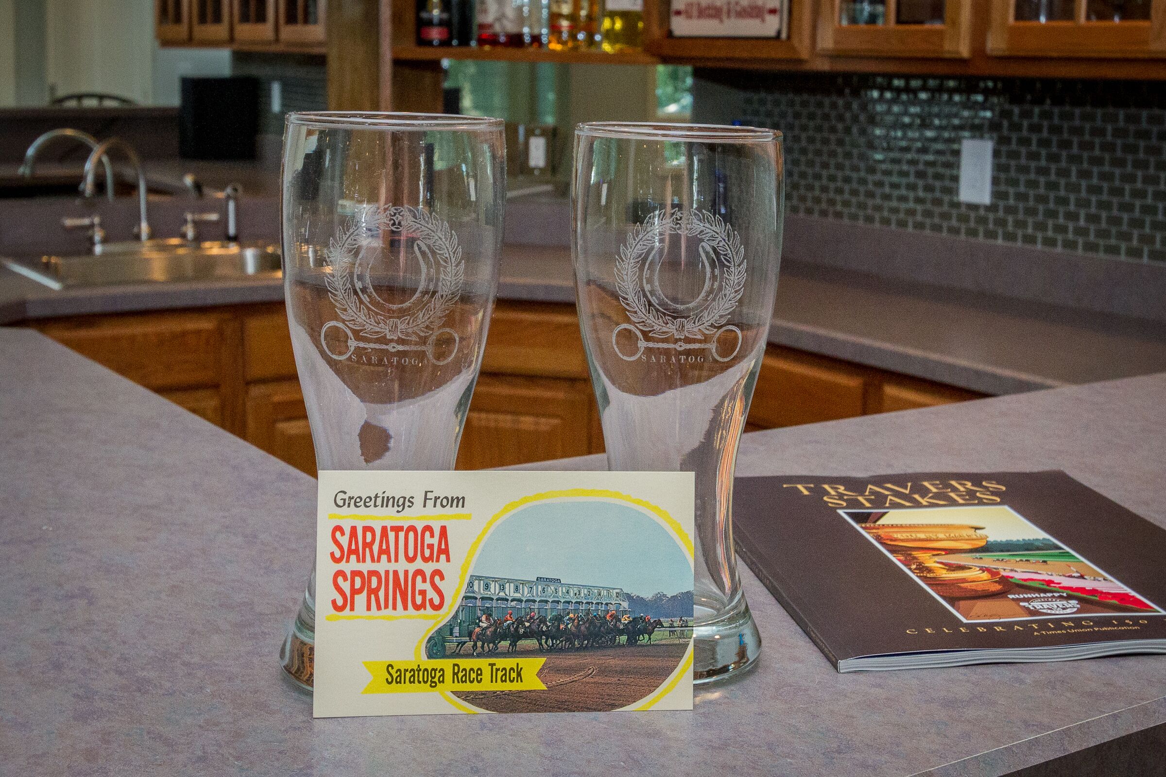 Two clear glasses with embossed logos on a bar counter, a card with 'Greetings From Saratoga Springs' and a race track image, and a book titled 'TRAVELING STAKES' with a horse racing scene on the cover.