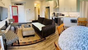 A compact living space with a sofa, ottoman, and a dining table. A kitchen area with modern appliances is visible in the background.