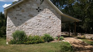 Ein Steinhaus mit Kruzifix an der Wand, umgeben von Grünpflanzen und einem Kiesweg.