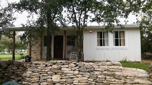 Ein weißes Haus mit einer Steinmauer, einer Veranda mit Stühlen und einem kleinen Fenster mit Vorhängen.