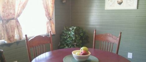 A dining table with a bowl of fruit, surrounded by wooden chairs, in a room with a green wall and a window with curtains.
