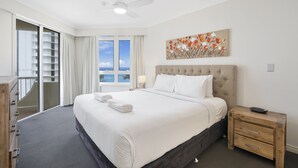 Une chambre d’hôtel avec un grand lit, un ventilateur de plafond, une fenêtre donnant sur un beau paysage, une table de chevet en bois et un tableau au mur.