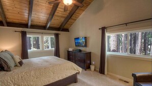 Une chambre avec un grand lit, un ventilateur de plafond, un téléviseur posé sur une commode et une fenêtre donnant sur une forêt.