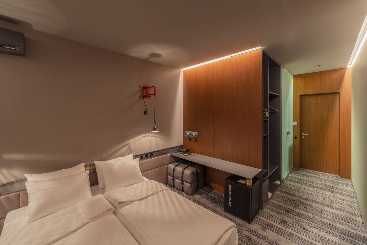 A hotel room with a bed, bedside table, lamp, wardrobe, and a small seating area.
