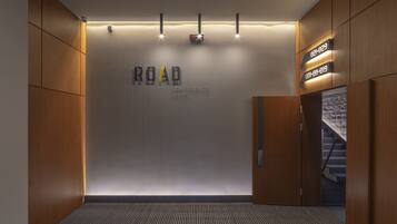 A modern hotel lobby with a 'ROAD CONFERENCE HOTEL' sign, a reception desk, and a staircase leading to the upper floors.