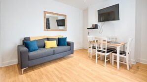 Ein modernes Wohnzimmer mit einem grauen Sofa, blauen und gelben Kissen, einem Holztisch und weißen Stühlen. Ein Flachbildfernseher ist über dem Sofa an der Wand befestigt.