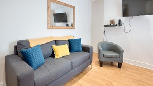 Ein modernes Wohnzimmer mit grauem Sofa, blauen und gelben Kissen, einem spiegelnden Wandbild mit Holzrahmen, einem grauen Sessel und einem fest montierten Flachbildfernseher.