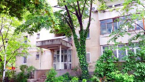 Un immeuble à plusieurs étages doté d’un balcon, entouré d’une végétation luxuriante et d’une clôture en métal.