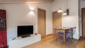 Un soggiorno moderno con una televisione a schermo piatto su un mobile bianco, porte in legno e un'area pranzo con sedie colorate.