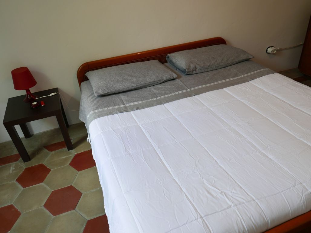 A neatly made bed with a white duvet and grey pillows, a small bedside table with a red lamp, and a hexagonal patterned floor.