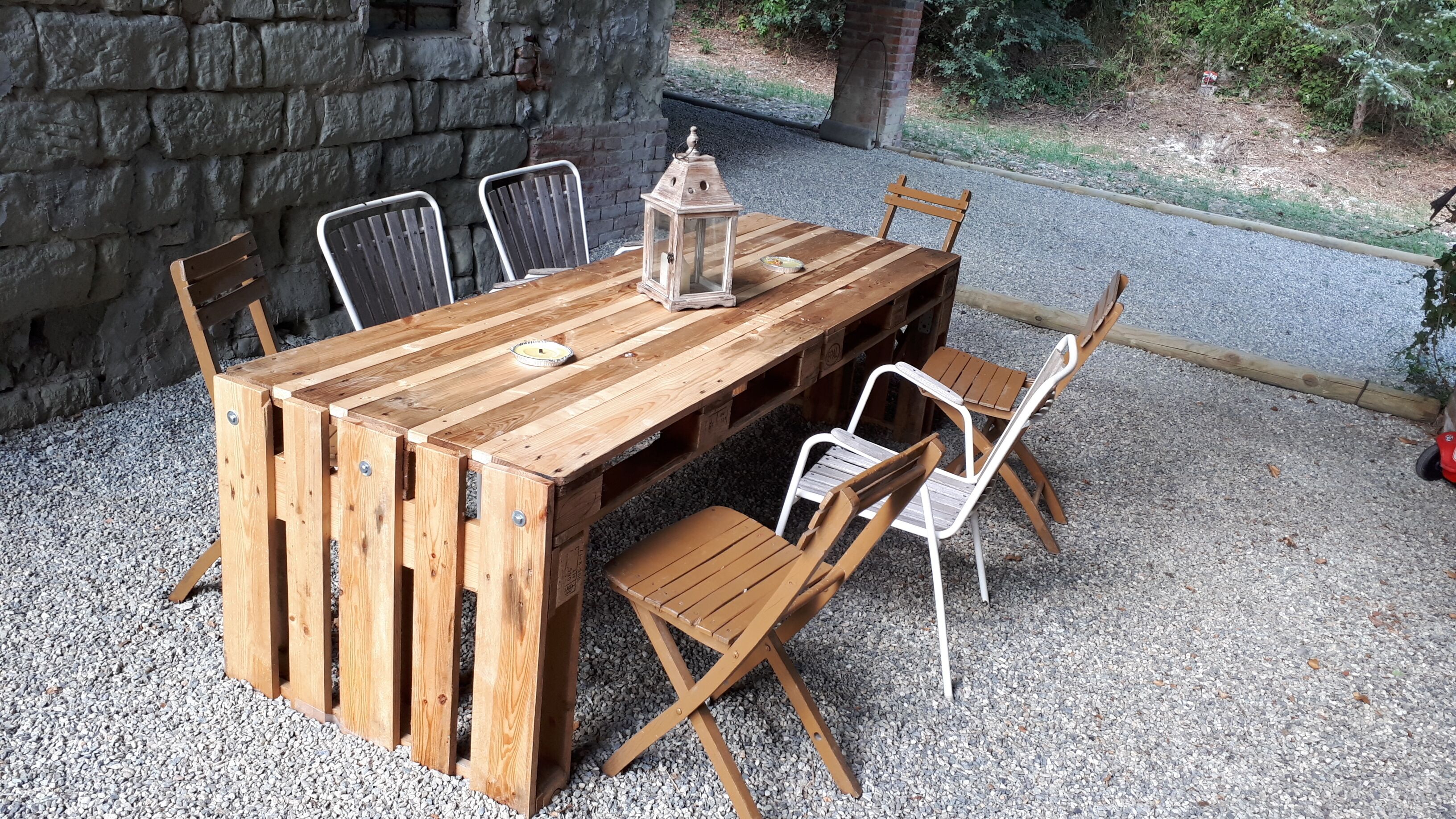 A rustic wooden table with chairs, a lantern, and a small plate on a gravel surface.