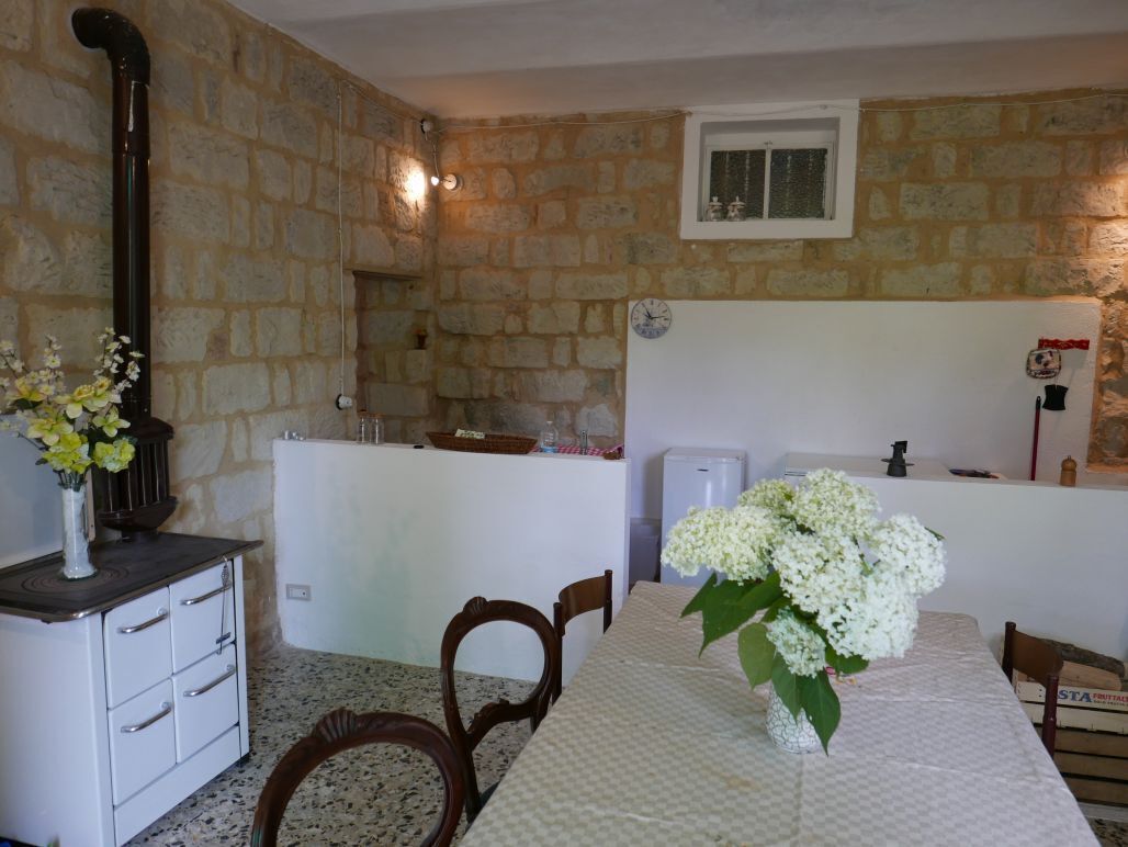 Habitació de paredats de pedra amb una taula blanca, cadires, un ram de flors, una cuina i una finestra.