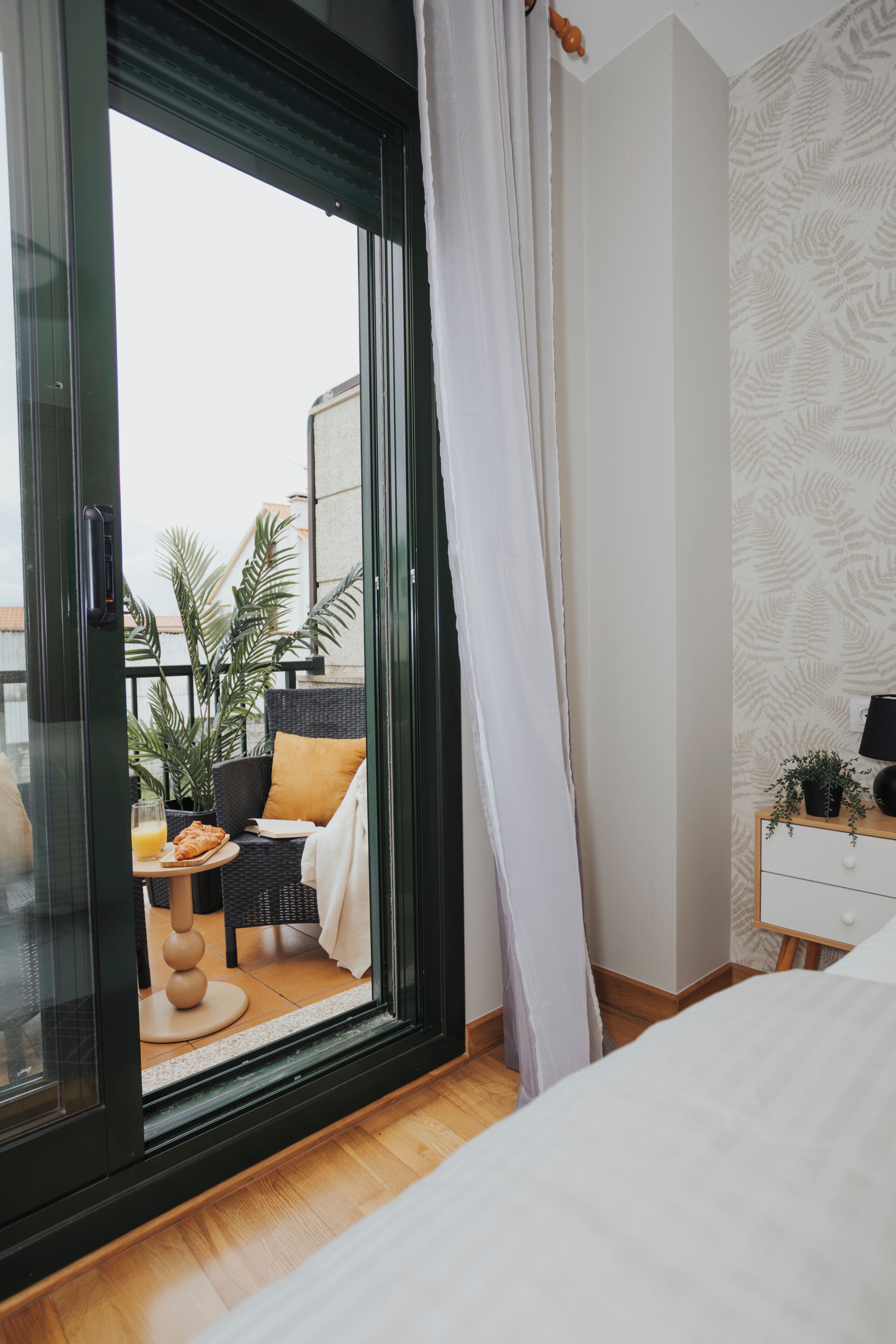 Un dormitorio con un ventanal grande, una cama, una mesita de noche con lámpara y vistas a un área de estar exterior con plantas.