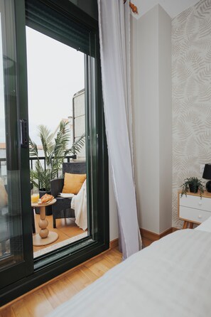 Quarto com uma janela ampla, uma cama, mesa de cabeceira com abajur e vista para uma área externa com mobiliário e plantas.
