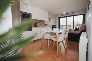 Een moderne keuken met witte kasten, een magnetron en een rode koffiezetapparaat. Er is een eettafel met stoelen en een balkon met uitzicht op de stad.