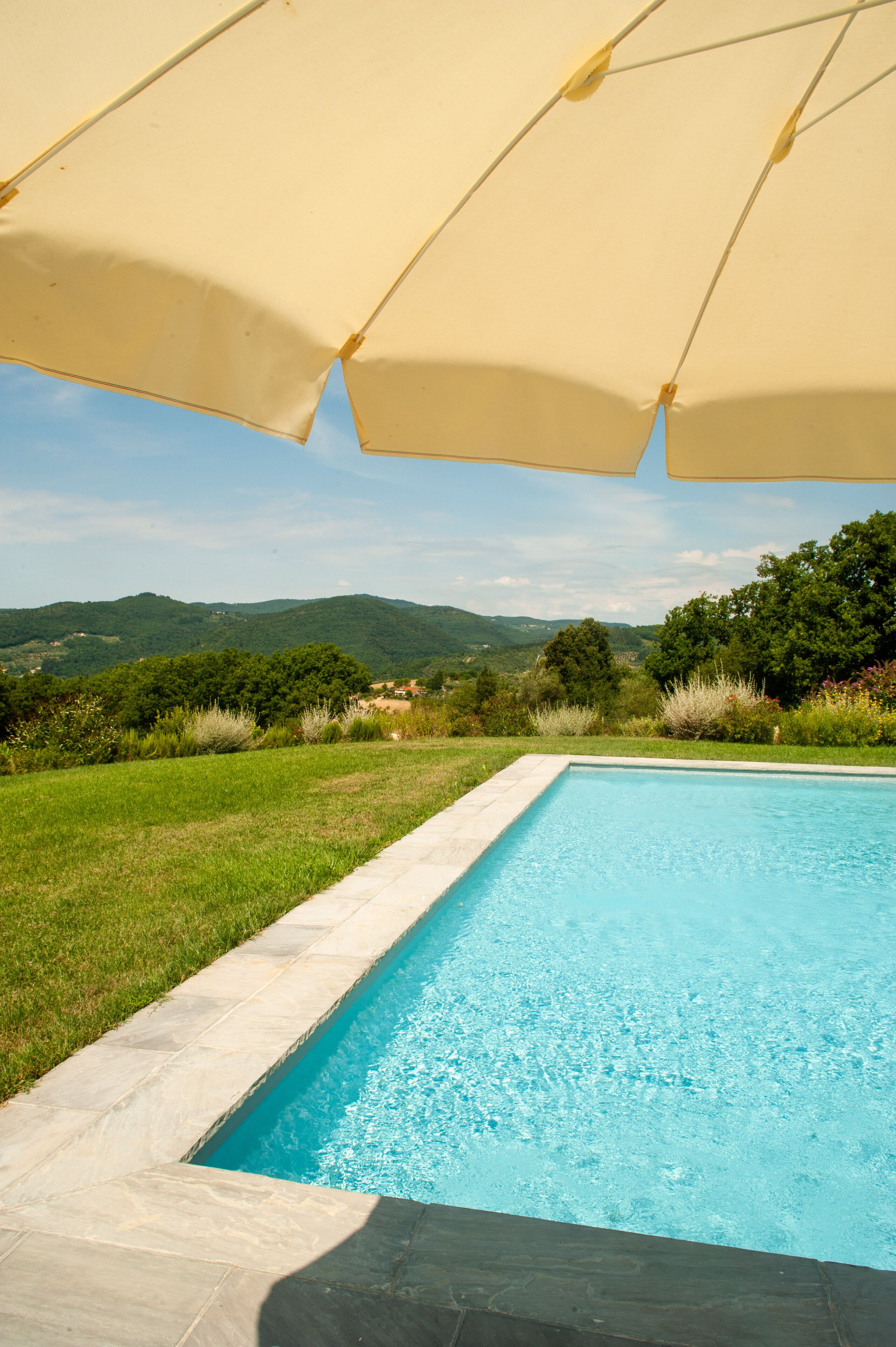 Una piscina con borde de piedra, rodeada de césped y árboles, con vistas a colinas al fondo.