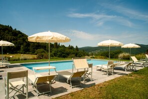 Un espace aménagé au bord de la piscine, avec des chaises longues et des parasols, entouré de verdure et de collines.