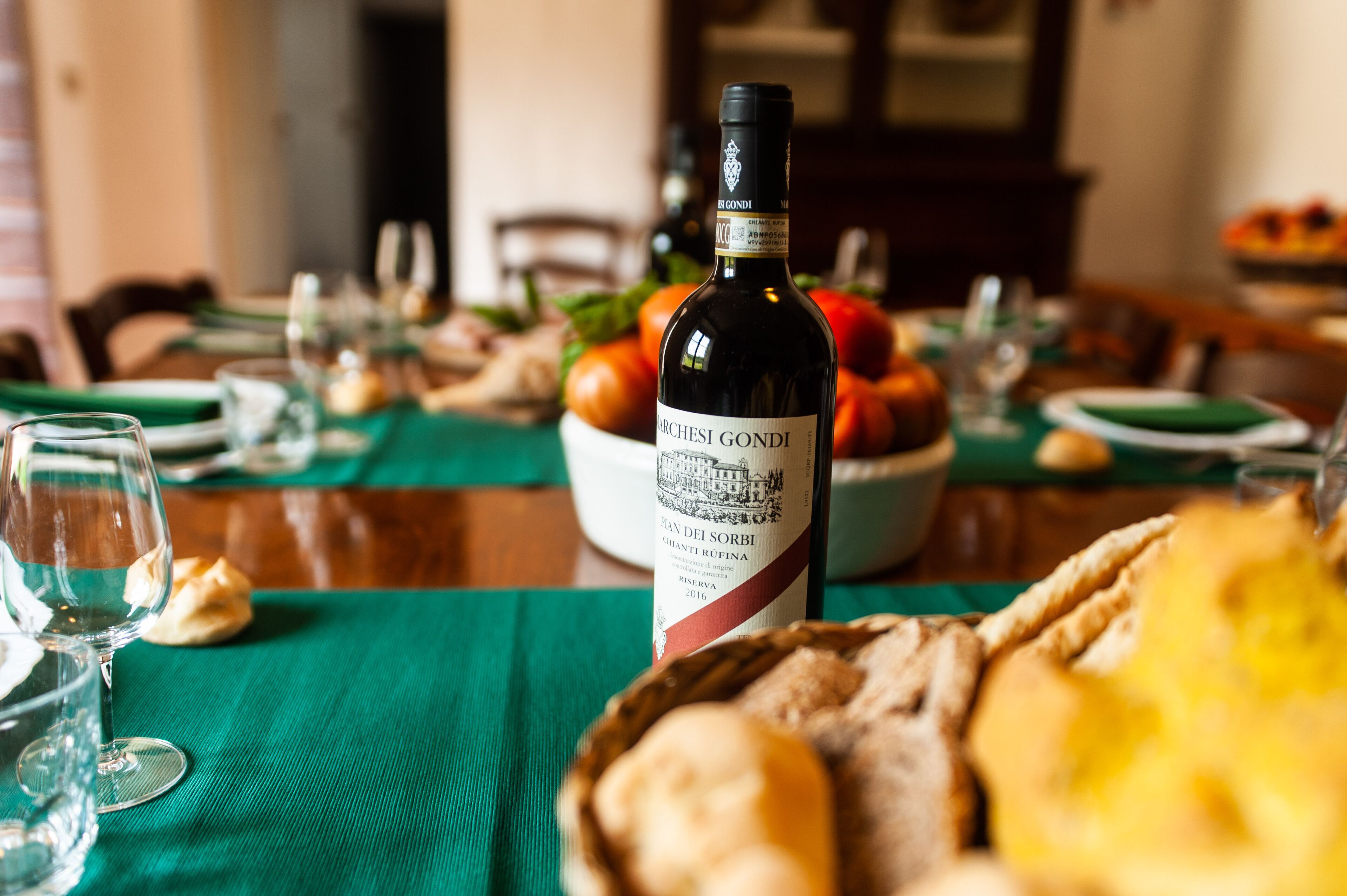 A dining table set with a bottle of wine, glasses, plates, and a bowl of fruit.