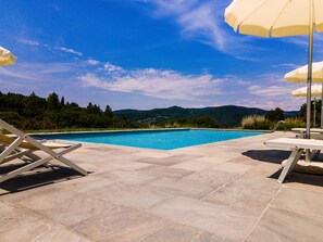 Una piscina con suelo de baldosas, sillones de descanso y sombrillas.