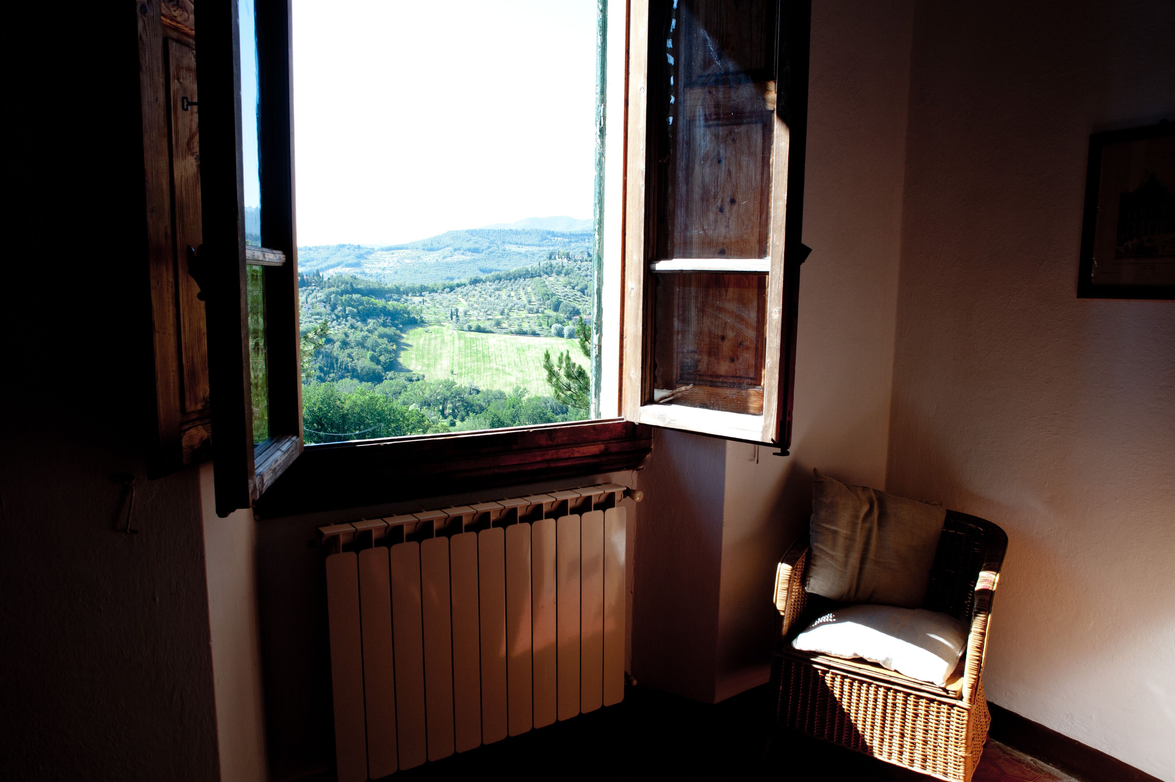 A room with a view of a green landscape through an open window, a wicker chair, and a radiator.