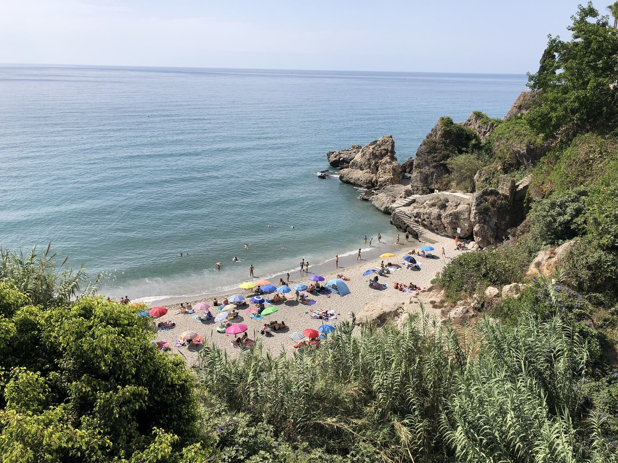 A beach with people sunbathing and swimming, surrounded by rocky cliffs and greenery.