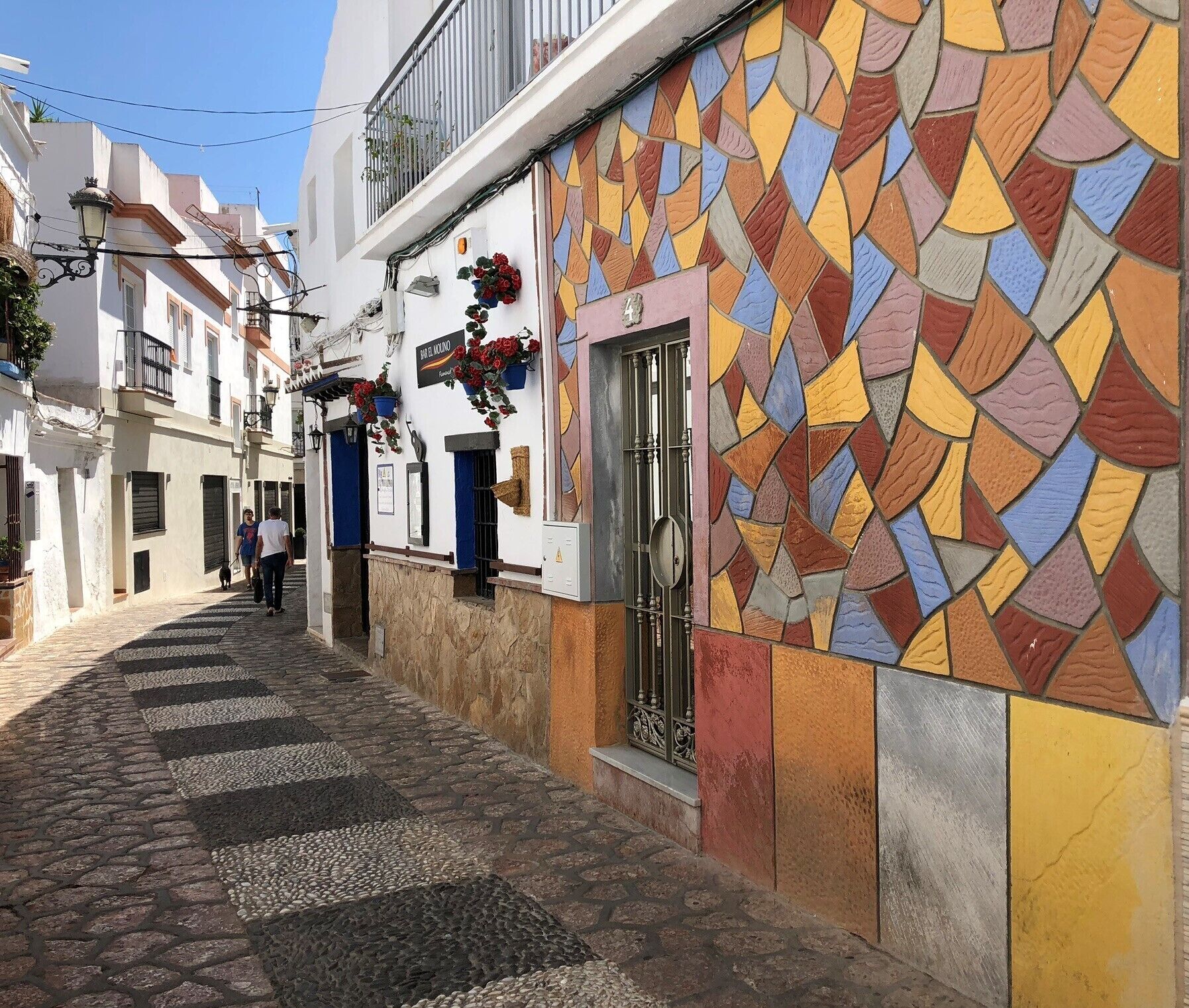 Una estrecha calle adoquinada flanqueada por edificios con coloridos mosaicos y un letrero que indica un restaurante.