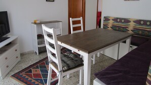 Ein Essbereich mit einem Holztisch und weißen Stühlen, einem Fernseher auf einem weißen Schrank und einem gemusterten Teppich.
