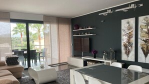 A modern living room with a sofa, a dining table, and chairs. There is a TV mounted on the wall, and a window with a view of palm trees. - Luxury chalet: private pool, close to the beach, W-Lan incl., quiet location, village within walking distance  (Cala Mendia)