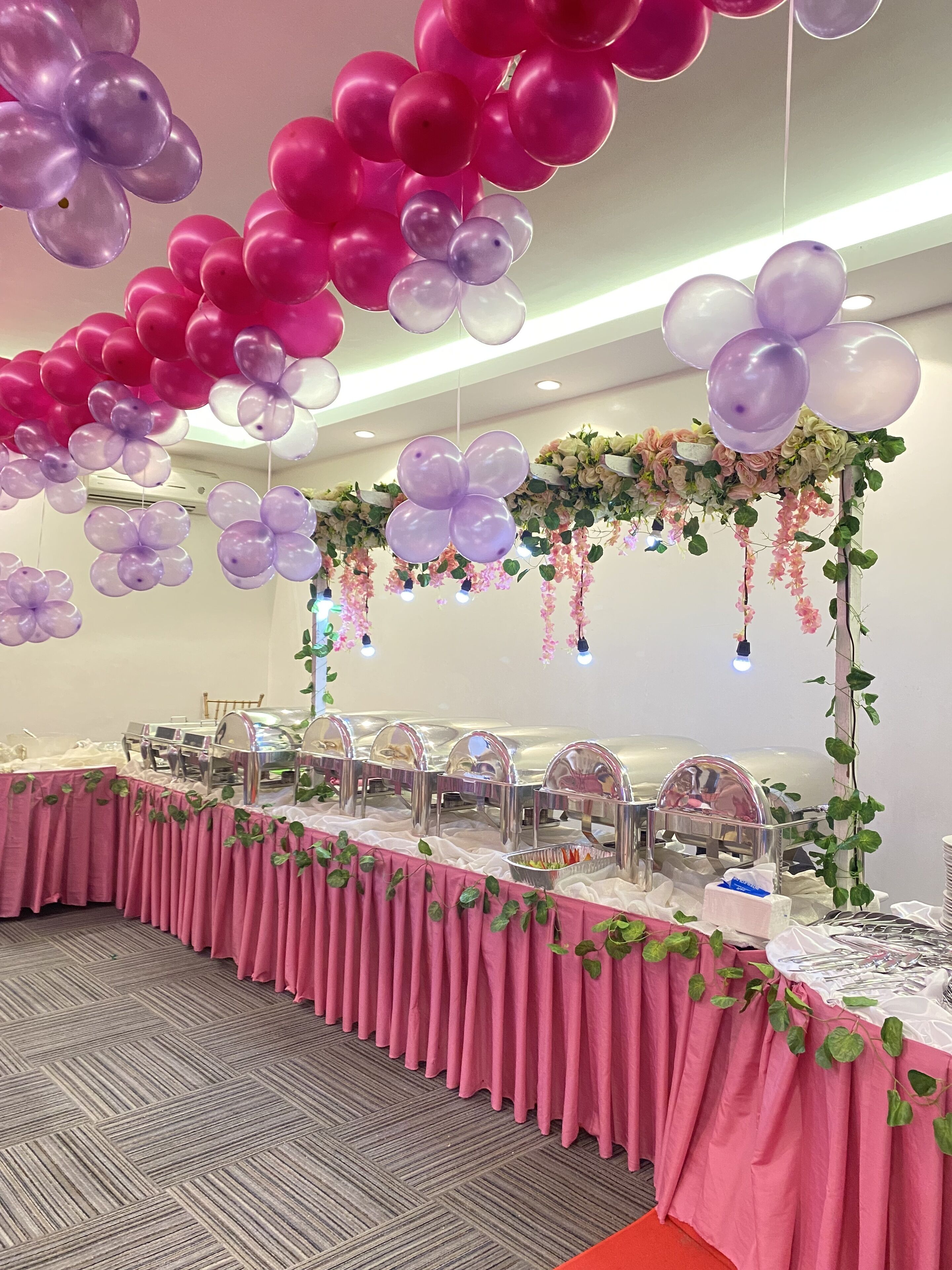Salão de banquetes decorado com balões rosa e roxos, flores e mesas de buffet.