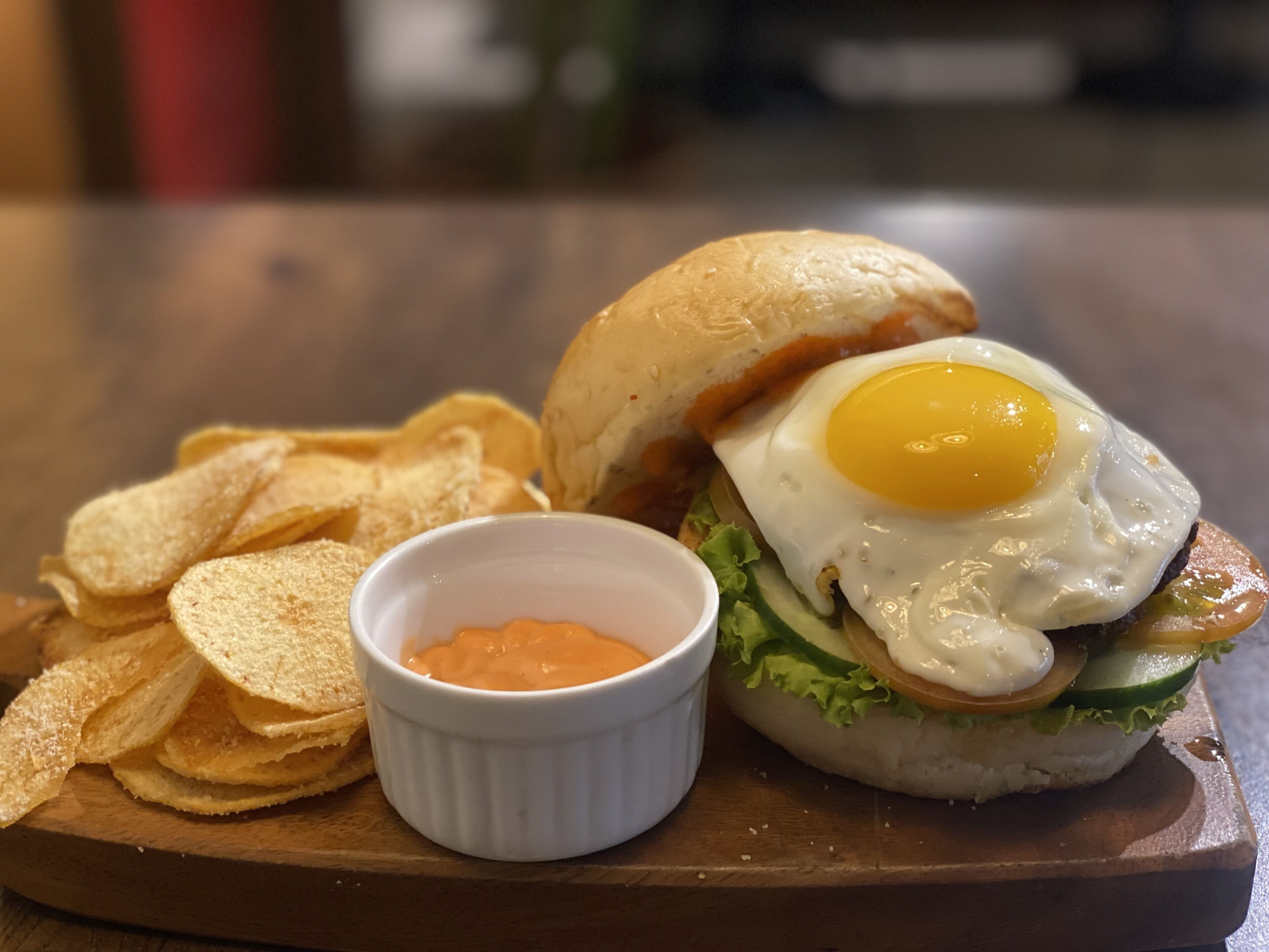 A sandwich with a fried egg, lettuce, tomato, and a bun, accompanied by chips and a small bowl of sauce on a wooden board.