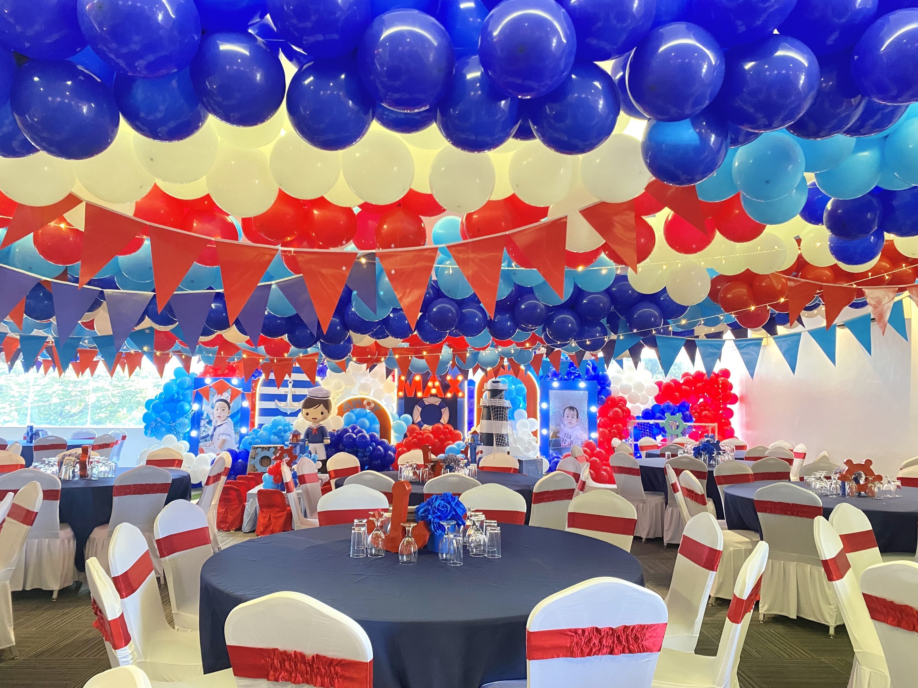 A banquet hall with round tables, white chairs with red sashes, and a ceiling adorned with blue and red balloons.