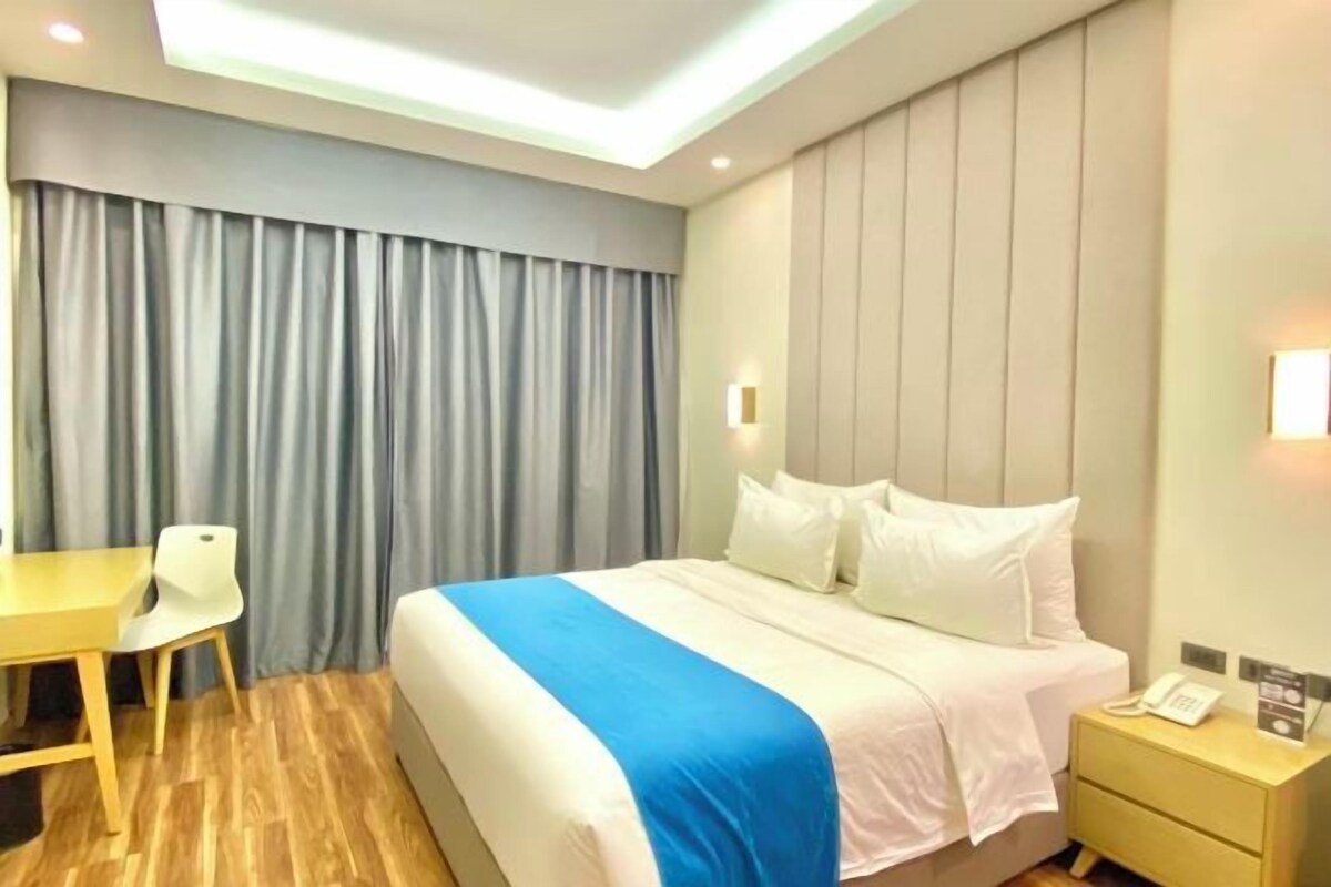 Habitación de hotel con cama, mesita de noche, lámpara y escritorio con silla.