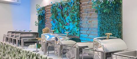 A buffet setup with stainless steel chafing dishes and a decorative wall with greenery.