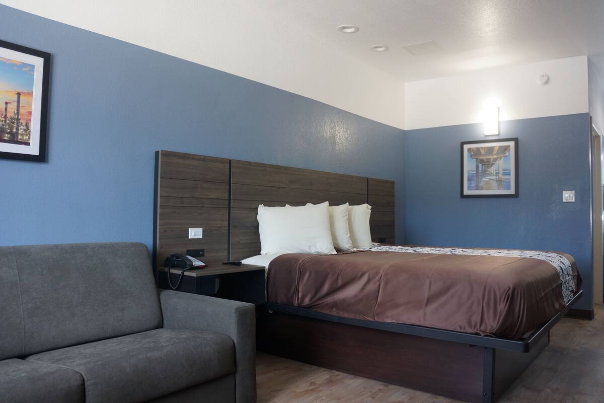 A hotel room with a bed, a gray armchair, a nightstand with a phone, and two framed pictures on the wall.