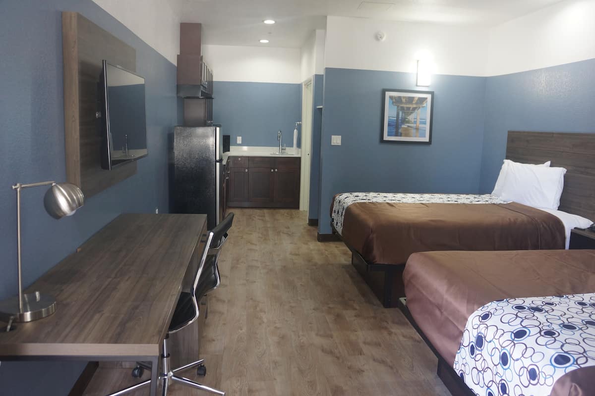 A compact hotel room with a wooden desk, two beds, a kitchenette, and a large mirror.