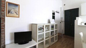 A room with a television on a white table, a cabinet with glass doors, a framed picture of Geneva airport, and a door with a coat hook. - Italianway - Bligny 64 (Milan)