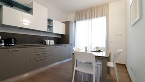 A modern kitchen with grey cabinets, a coffee machine, and a dining area with white chairs and a table. - Italianway - Gambara 7-6 (Milan)