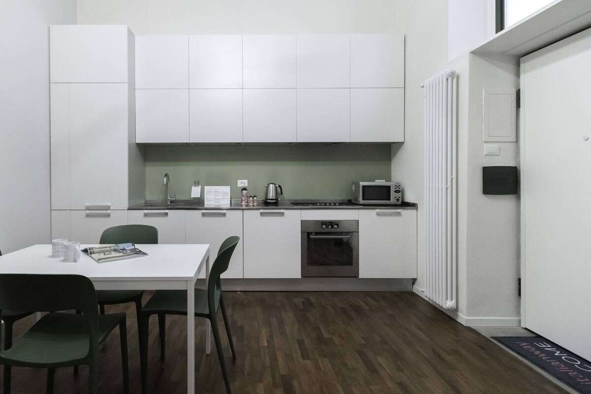 Une cuisine compacte avec des meubles blancs, un four à micro-ondes, une bouilloire et un petit four. On y trouve une table à manger avec quatre chaises et quelques objets posés dessus.