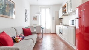 Eine moderne Küche mit weißen Schränken und einem roten Kühlschrank, ein graues Sofa mit roten Kissen sowie eine Essecke mit einem weißen Tisch und Stühlen.